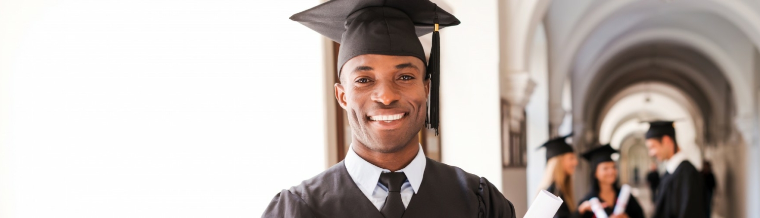 A graduate in cap and gown smiles while holding a diploma, standing in a hallway with other graduates in the background.